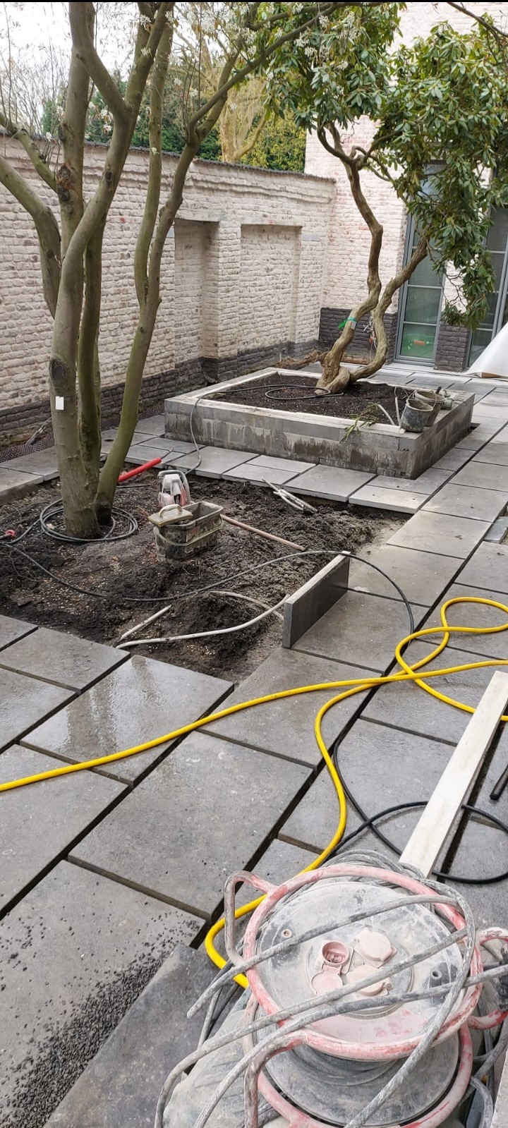 Układanie kostki brukowej wokół drzewa na dziedzińcu. Widoczne narzędzia i kable. Częściowo ułożona kostka tworzy ścieżkę i obrzeże rabaty. Ceglany mur w tle.