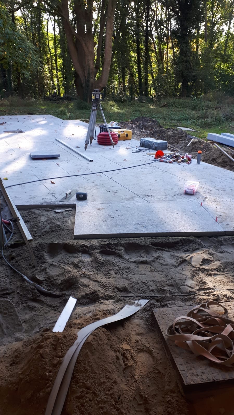 Układanie jasnoszarej kostki brukowej na piasku, widoczny niwelator laserowy, narzędzia i częściowo ułożona nawierzchnia na tle drzew w słoneczny dzień.