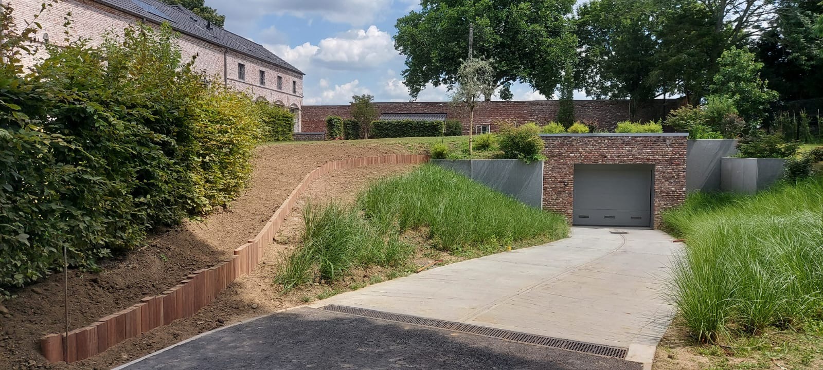 Ukształtowany teren z drewnianym obrzeżem i ozdobnymi trawami przy podjeździe do garażu w nowoczesnym stylu. Widok na budynek z cegły i zieleń.