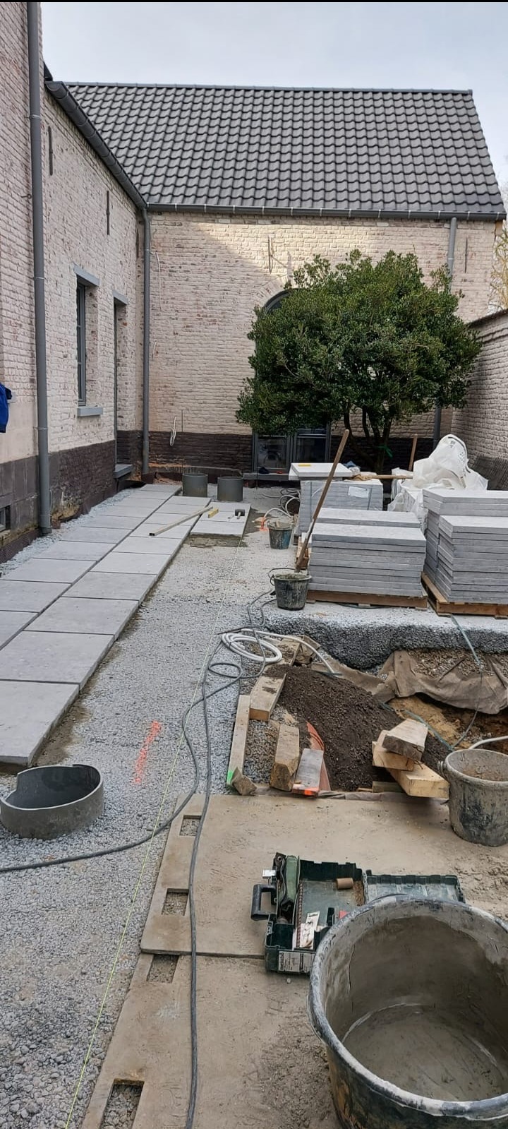 Układanie kwadratowych płyt chodnikowych na żwirowym podłożu wzdłuż ceglanego budynku. Widoczne narzędzia, wiaderka i stos płyt. Prace brukarskie w toku.