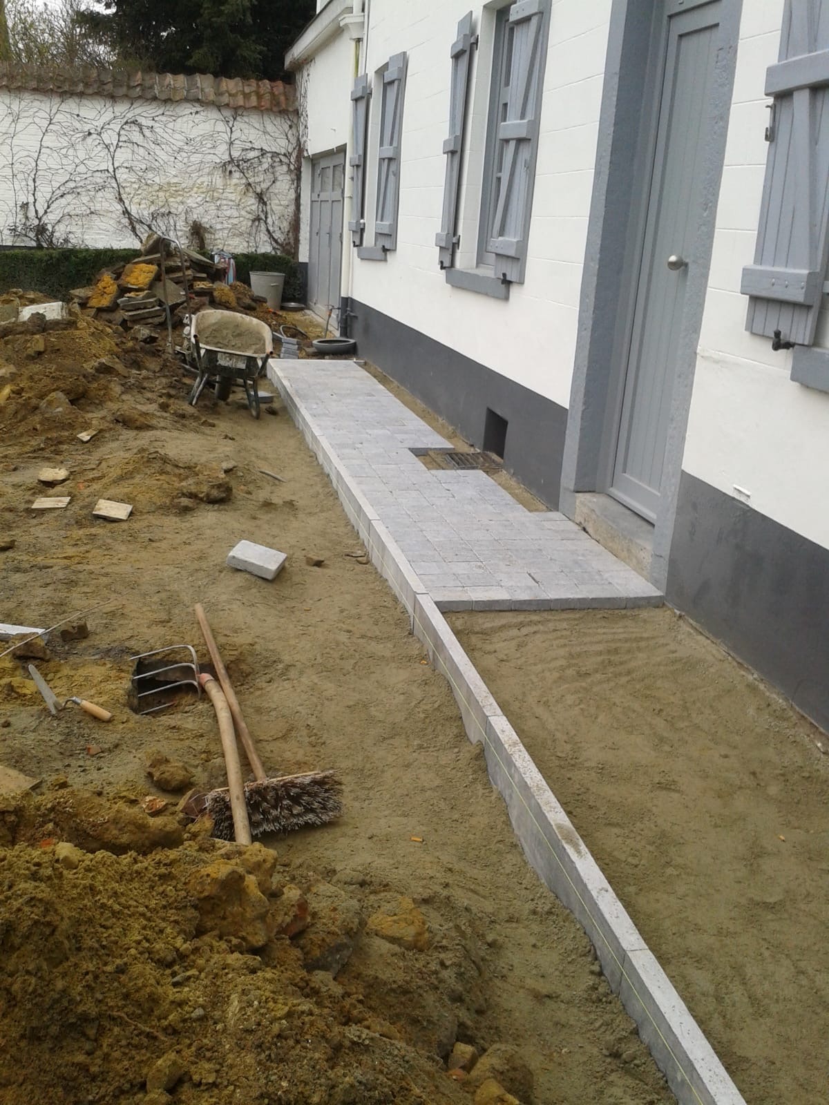 Układanie szarej kostki brukowej wzdłuż białego budynku z szarymi okiennicami. Widoczne narzędzia i piasek. Częściowo ukończony chodnik z kostki.