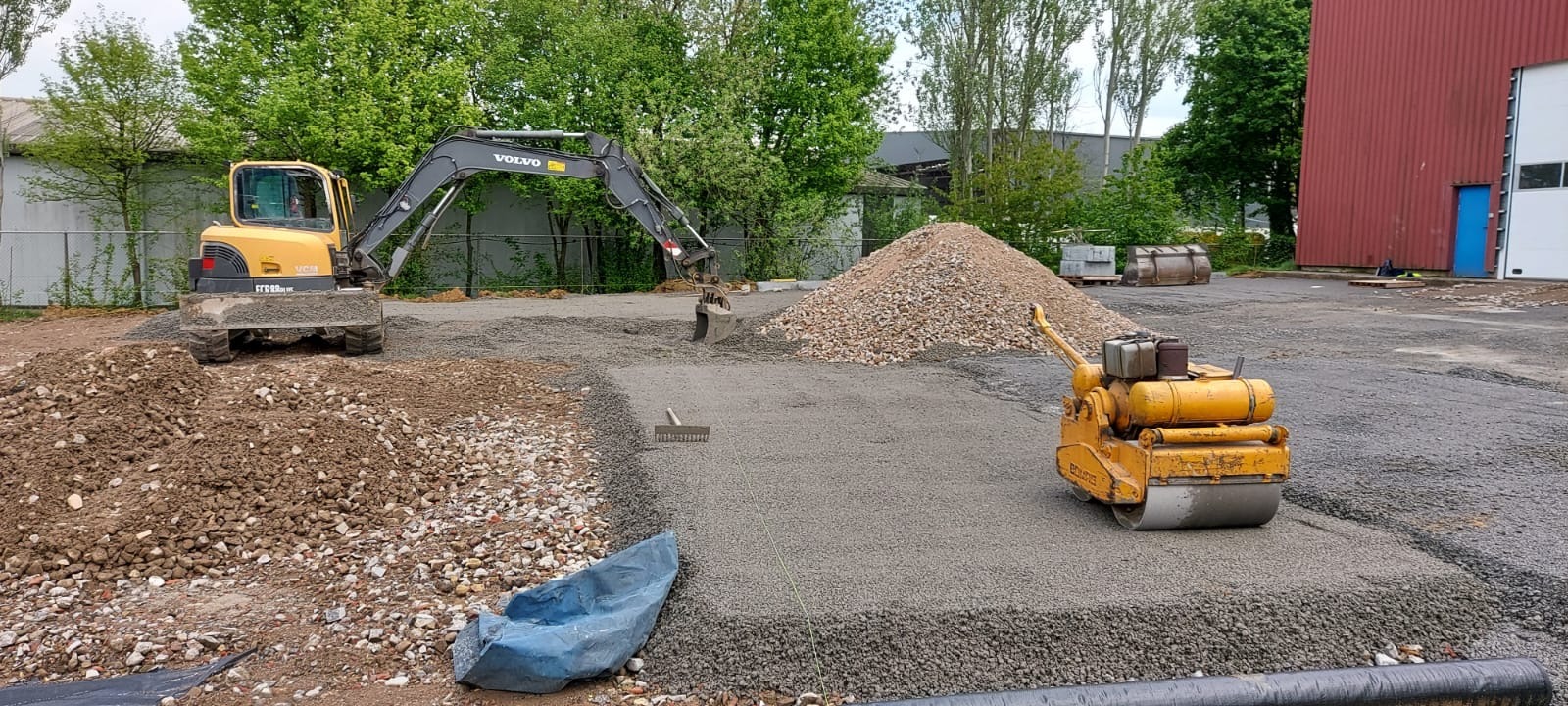 Żółty walec i koparka Volvo na placu budowy z przygotowanym podłożem z kruszywa. W tle sterta gruzu i budynek z czerwonej blachy.