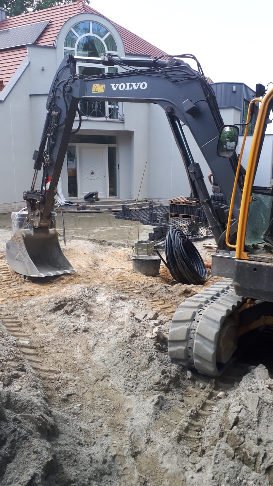 Częściowy widok koparki Volvo na tle domu z kostką brukową. Etap przygotowania terenu pod układanie kostki. Widoczne ślady opon i piasek.