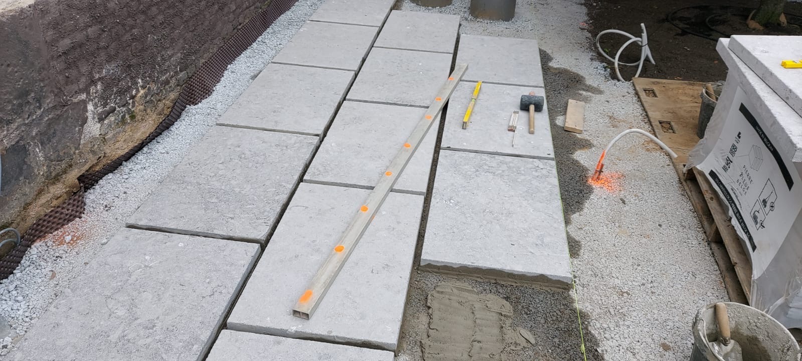 Układanie jasnoszarej kostki brukowej na podsypce. Widoczny poziomica, młotek i inne narzędzia. Etap prac brukarskich, przygotowanie do fugowania.