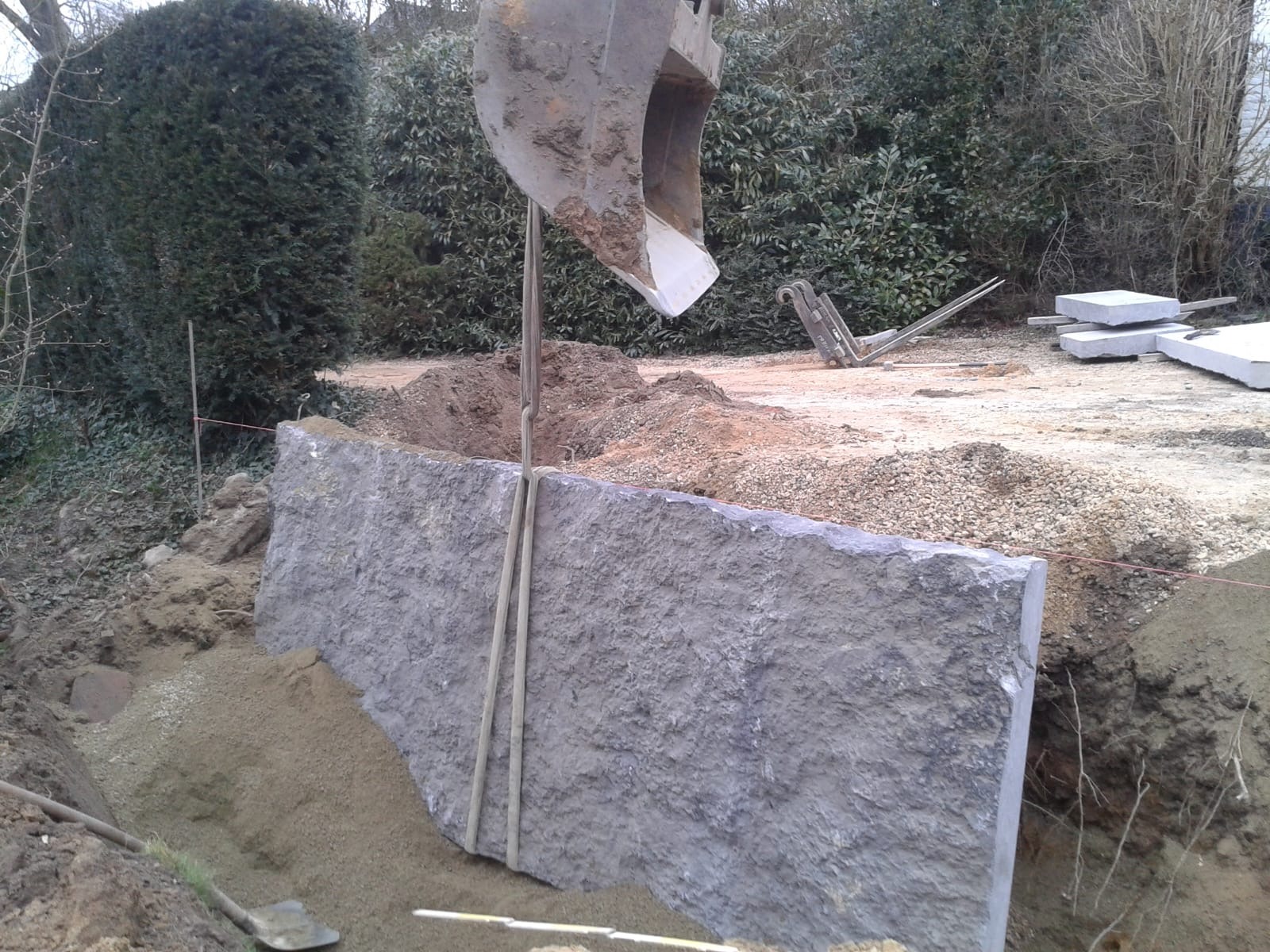 Montaż dużego, szarego bloku skalnego za pomocą koparki na terenie budowy. W tle widoczne krzewy i narzędzia.