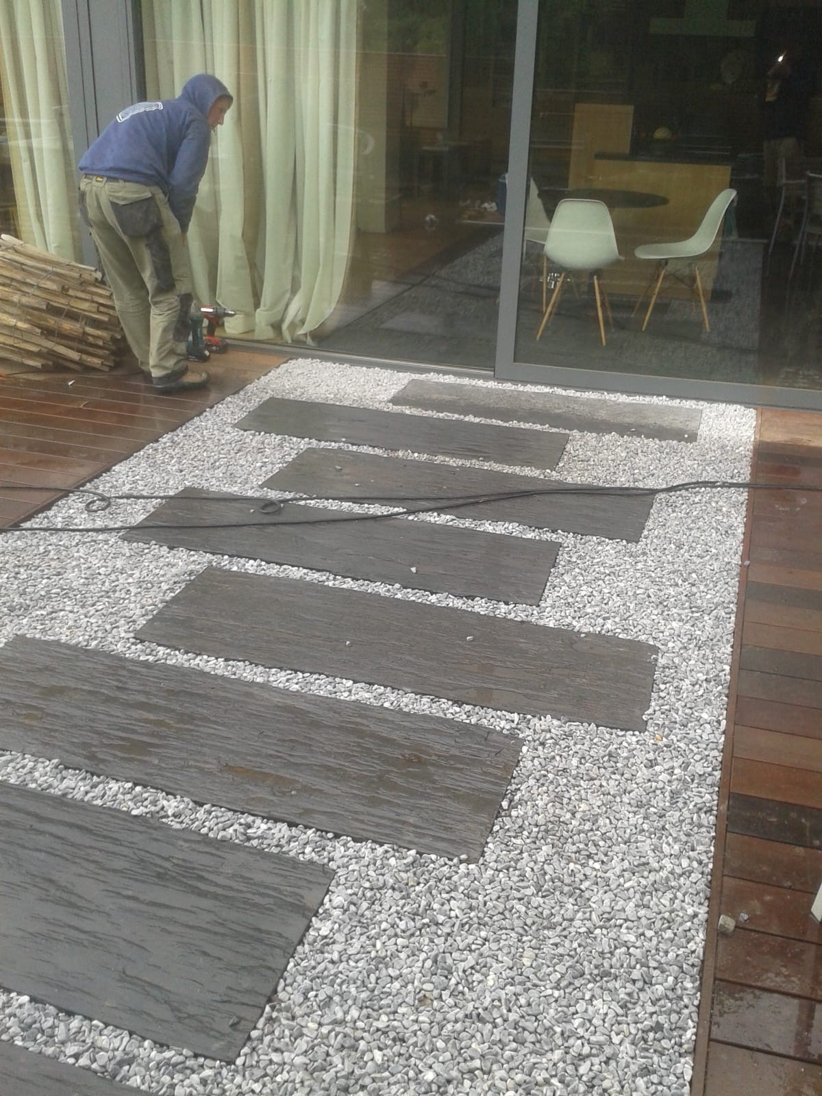 Ścieżka z płyt imitujących drewno, otoczona białym żwirem, prowadząca wzdłuż tarasu. Pracownik w trakcie montażu. Widoczne narzędzia i materiały.