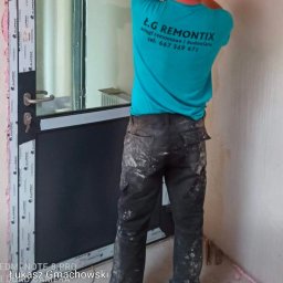 ŁG.REMONTIX - Pracownik w turkusowej koszulce z logo firmy montuje okno z czarną ramą. Widoczne pianka montażowa i narzędzia. W tle jasne ściany w trakcie remontu.