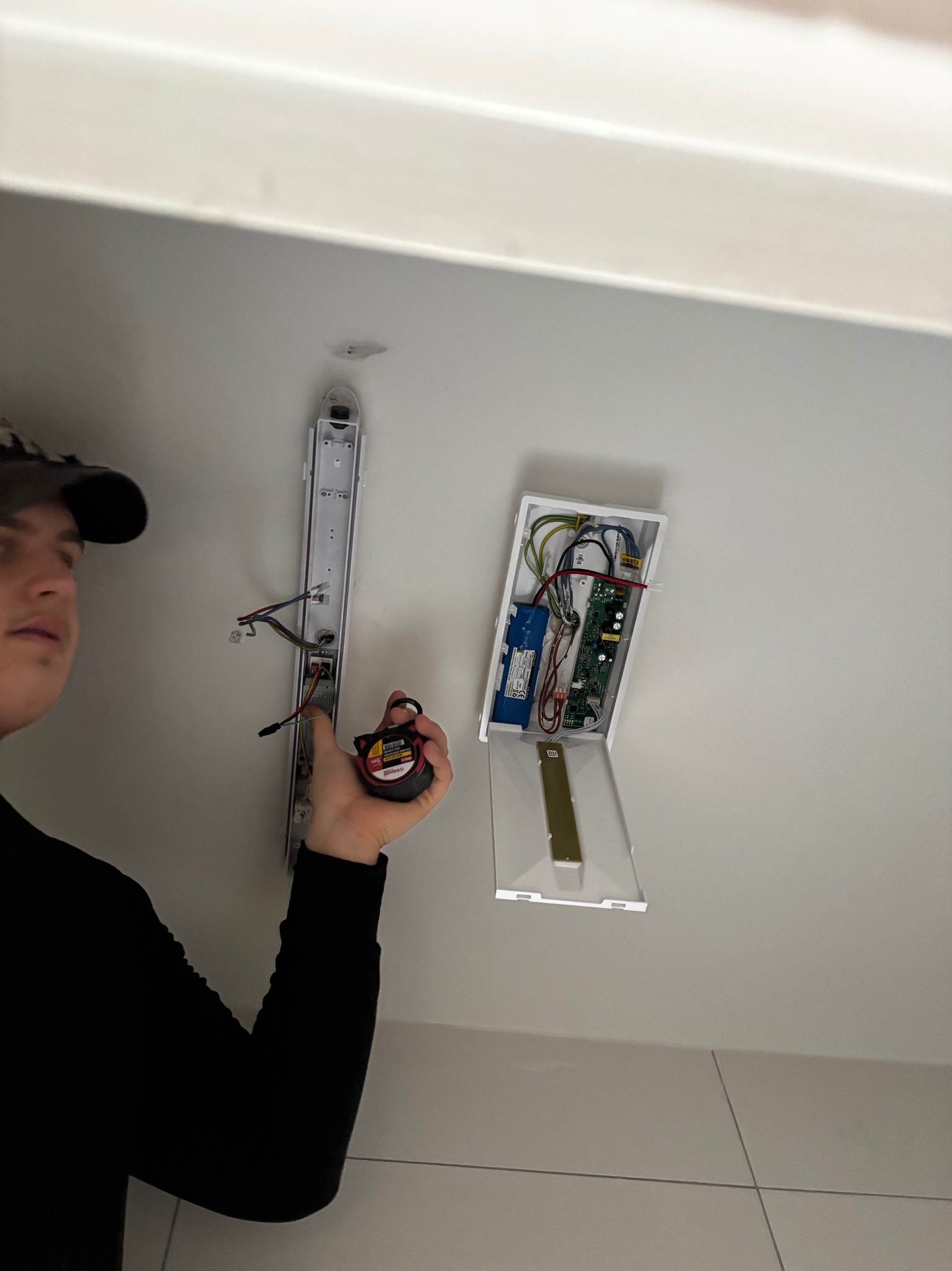 Elektryk mierzy odległość taśmą mierniczą do listwy oświetleniowej i otwartej skrzynki z elektroniką na suficie. Widoczne przewody i płytka drukowana.