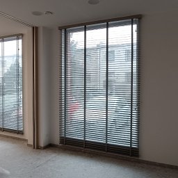 PRODOMUS Przemysław Jakubowski - Dwa okna z zamontowanymi żaluzjami drewnianymi w kolorze szarym. Widok na zewnątrz przez okna. Częściowo widoczne meble w pomieszczeniu.