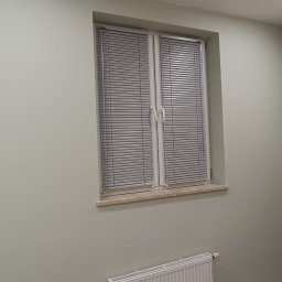 PRODOMUS Przemysław Jakubowski - Jasne, aluminiowe żaluzje poziome zamontowane w oknie. Widoczny biały grzejnik pod oknem, listwa z gniazdkami i narzędzia na podłodze zabezpieczonej folią.