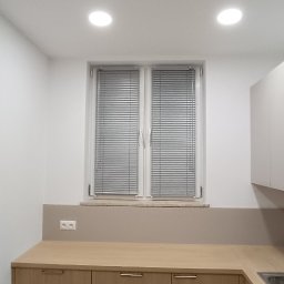 PRODOMUS Przemysław Jakubowski - Kuchnia z drewnianymi szafkami i aluminiowymi żaluzjami w oknie. Minimalistyczny design, jasne wnętrze, widoczne gniazdko elektryczne nad blatem.