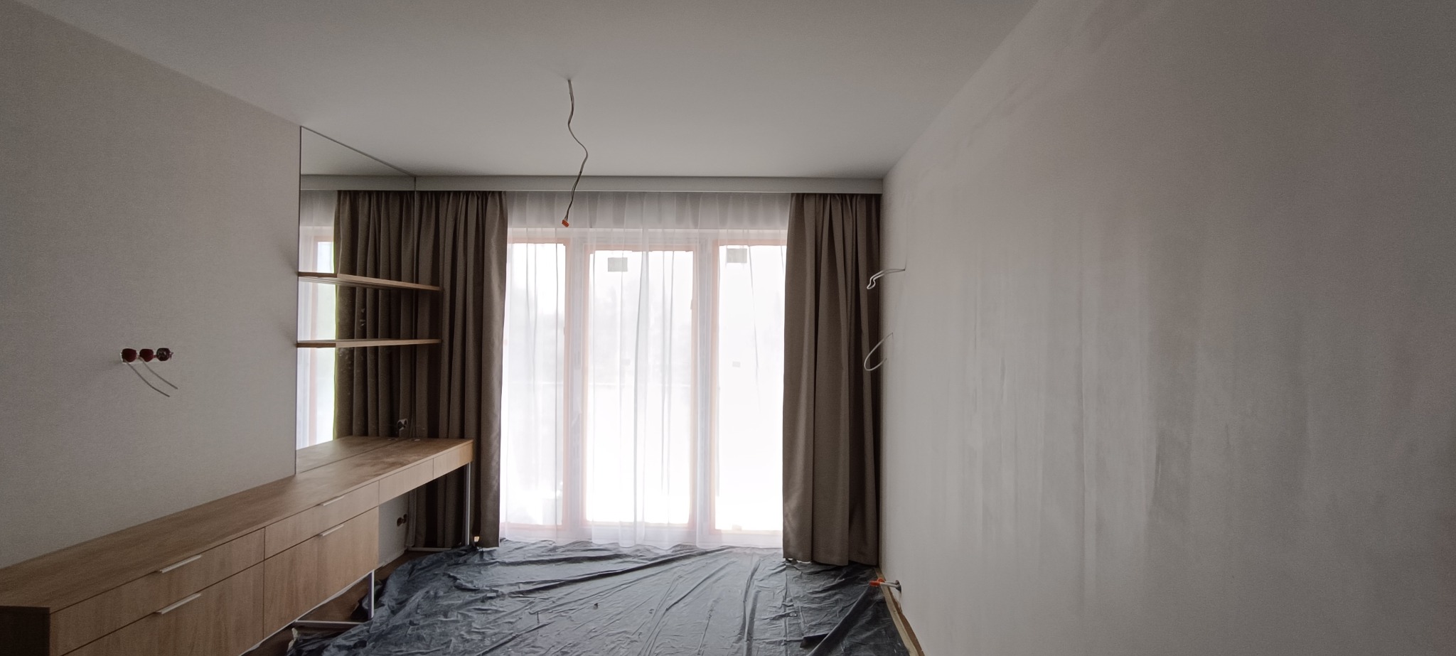Pokój w trakcie wykańczania: zasłony w oknach, meble z jasnego drewna, ściany przygotowane do malowania, podłoga zabezpieczona folią. Widoczne kable elektryczne.