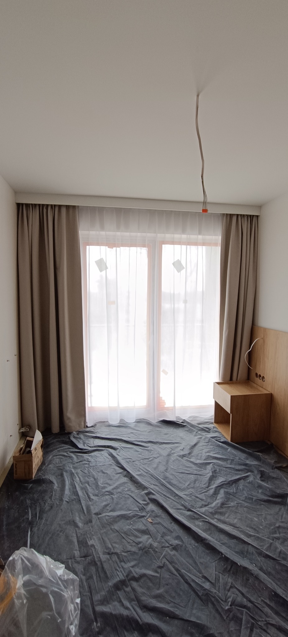 Pokój z zasłoniętymi oknami i zabezpieczoną podłogą folią malarską. Widoczne kable wystające z sufitu i drewniana szafka nocna. Przygotowanie do prac wykończeniowych.
