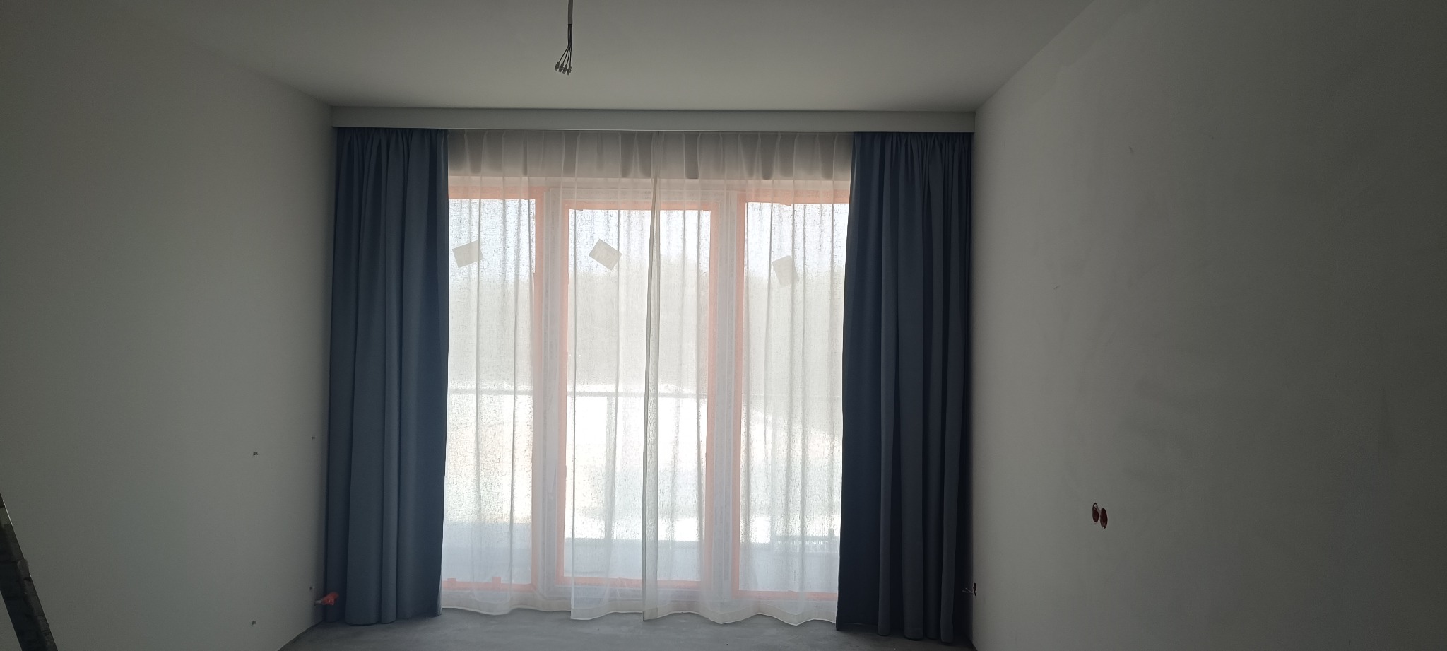 Jasne wnętrze z oknem, na którym wiszą długie, szare zasłony po bokach i delikatne, białe firanki na środku. Widoczne niedokończone ściany i wystające kable.