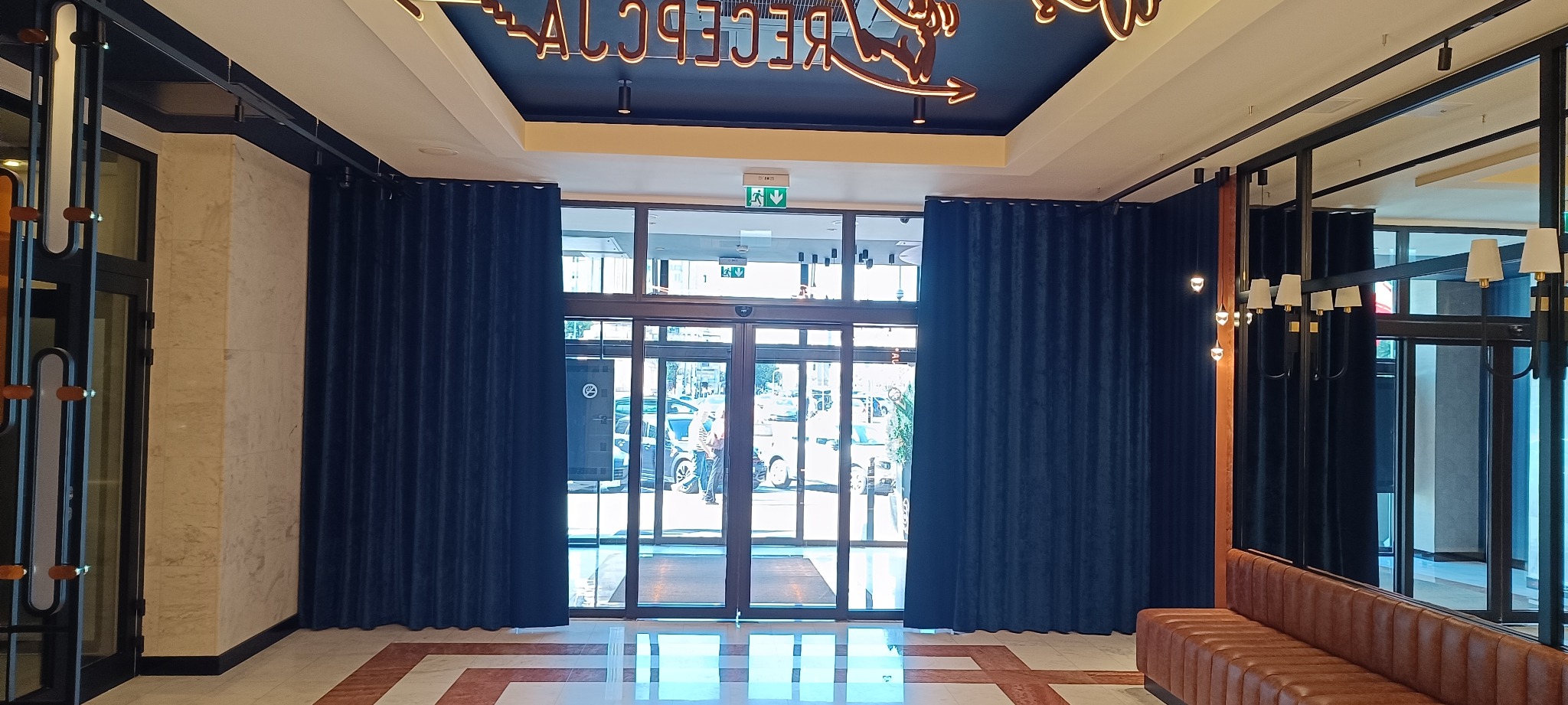 Eleganckie, granatowe zasłony w holu recepcyjnym w Poznaniu. Widoczne szklane drzwi wejściowe i fragment sofy. Oświetlenie sufitowe dodaje luksusowego charakteru.