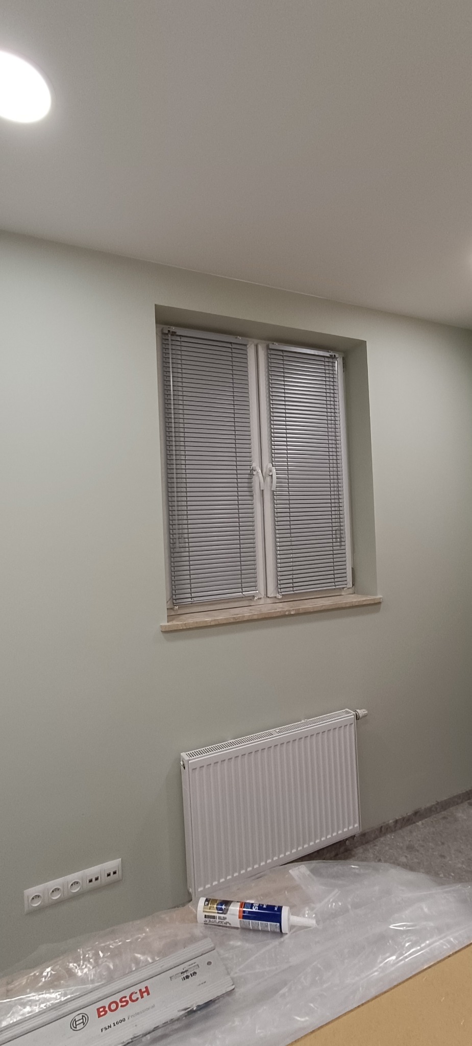 Jasne, aluminiowe żaluzje poziome zamontowane w oknie. Widoczny biały grzejnik pod oknem, listwa z gniazdkami i narzędzia na podłodze zabezpieczonej folią.