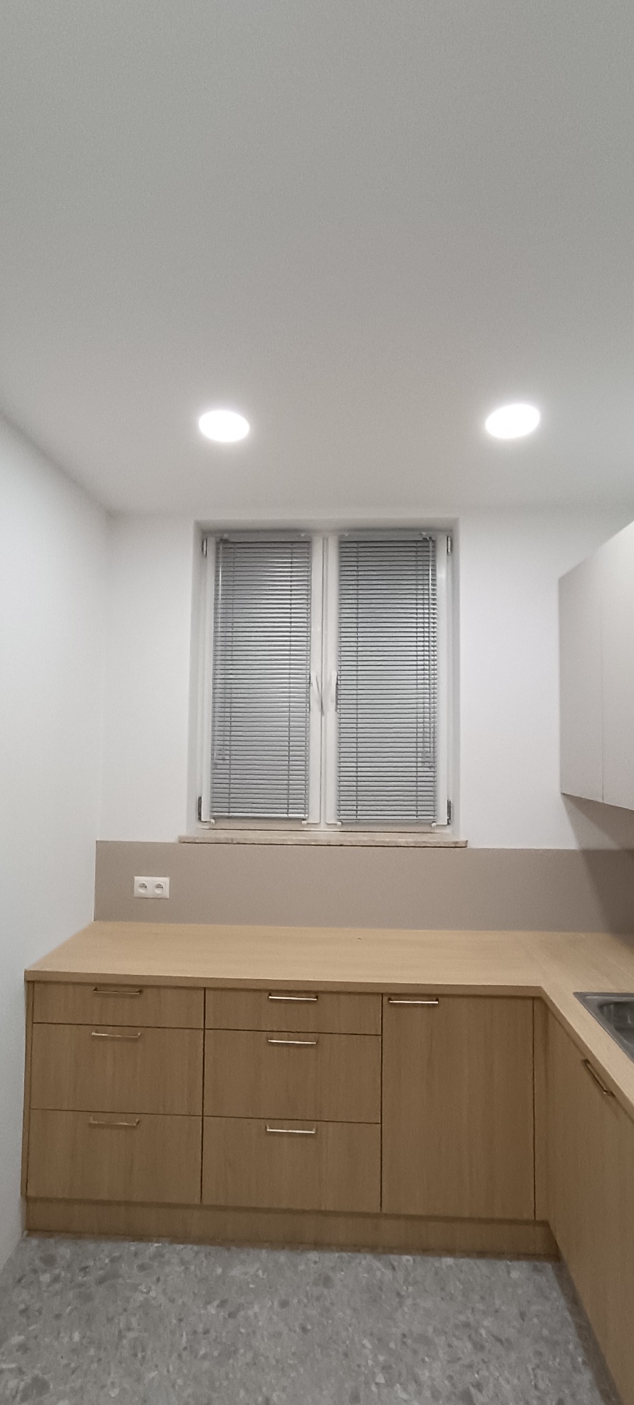 Kuchnia z drewnianymi szafkami i aluminiowymi żaluzjami w oknie. Minimalistyczny design, jasne wnętrze, widoczne gniazdko elektryczne nad blatem.