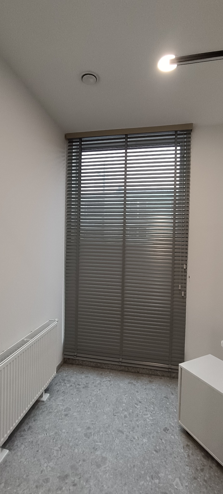 Szare poziome osłony okienne w pokoju z grzejnikiem i szafką. Widoczne sznurki do regulacji. Minimalistyczny wystrój wnętrza, neutralna kolorystyka.