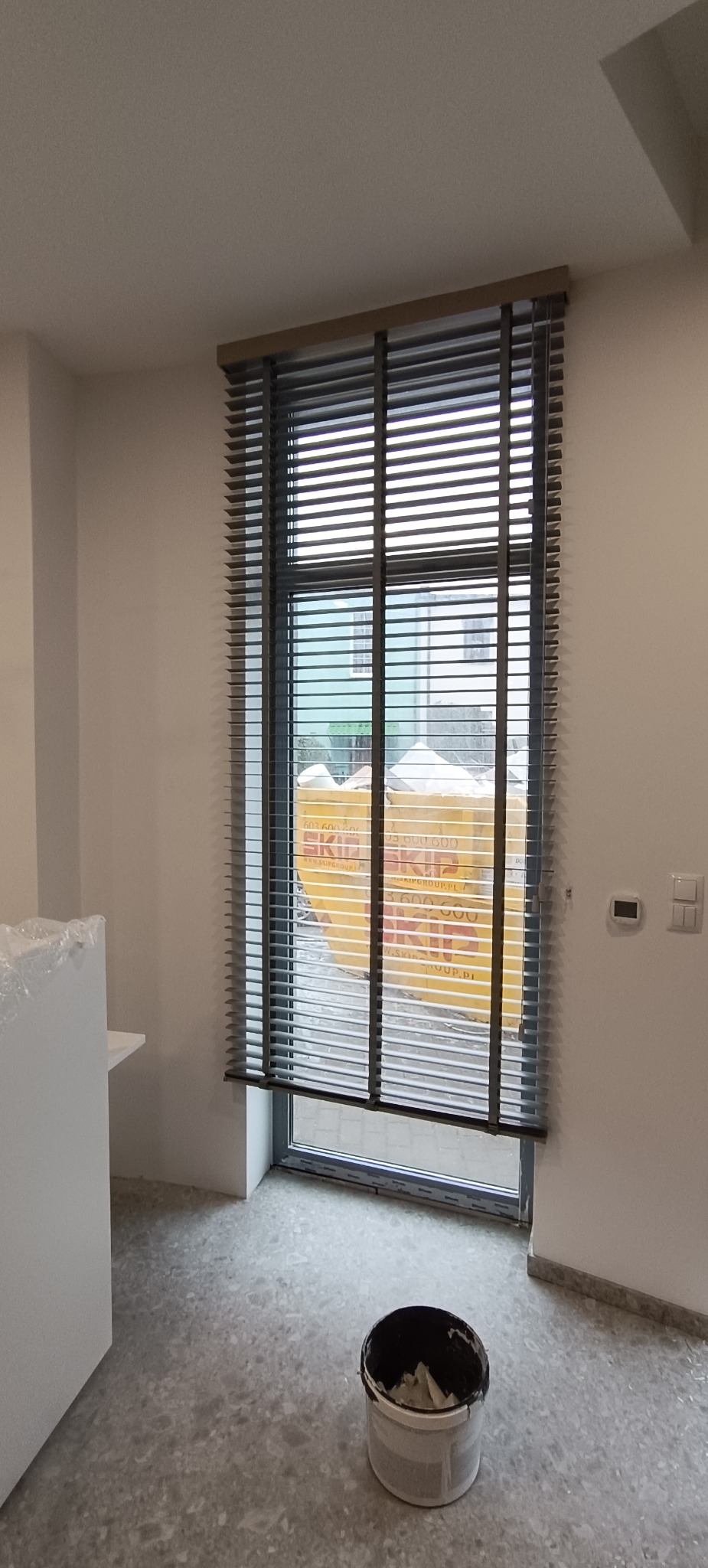 Zamknięte, poziome żaluzje w oknie, widok z wnętrza. Na podłodze wiadro z resztkami farby. W tle widoczne kartony za oknem.