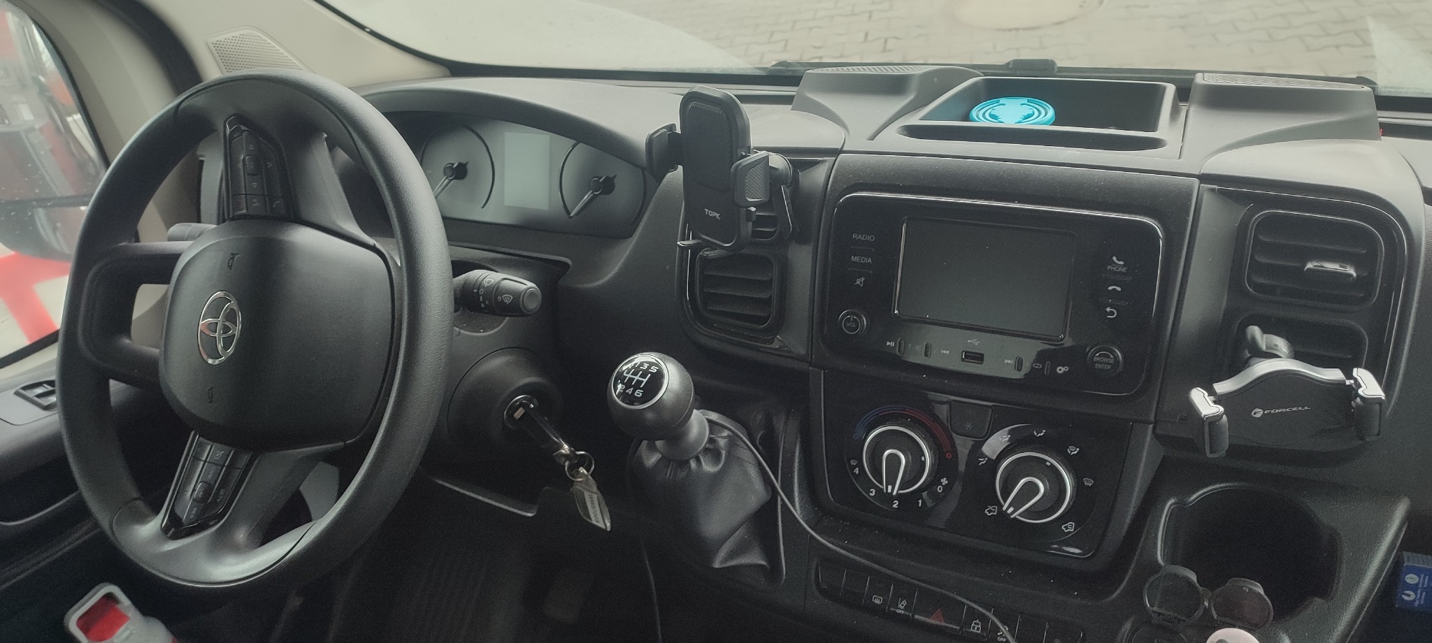 Wnętrze kabiny busa z kierownicą po lewej stronie, widoczny panel sterowania, dźwignia zmiany biegów i uchwyty na telefon. Kokpit w kolorze czarnym.