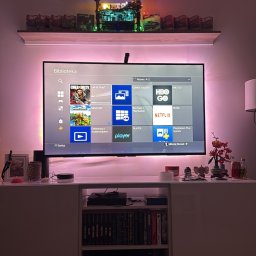 Mikołaj Nowak - Telewizor z podświetleniem LED na szafce RTV, nad nim półka z dekoracjami i zabawkami. Całość w stonowanej kolorystyce, oświetlona ciepłym światłem.