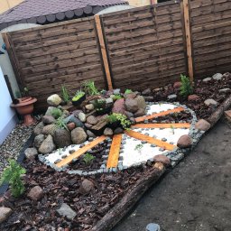 Mikołaj Nowak - Ogród z dekoracyjnym układem kamieni, drewna i białego żwiru, tworzącym wzór koła z promienistymi ścieżkami. Tło stanowi drewniany płot i fragment budynku.