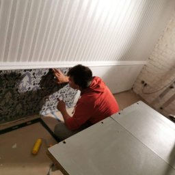 Mikołaj Nowak - Pracownik w czerwonej bluzie przykleja wzorzystą tapetę na ścianie pod skośnym sufitem. Widoczny wałek malarski i stół roboczy. Część ściany wykończona w białe pasy.