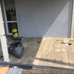 Mikołaj Nowak - Układanie drewnopodobnych płytek na tarasie. Widoczne poziomica, wiadro z zaprawą i narzędzia. Częściowo ułożona podłoga z płytek. Na zewnątrz, w słoneczny dzień.