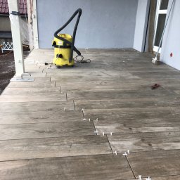 Mikołaj Nowak - Układanie drewnopodobnych płytek na tarasie. Widoczny odkurzacz przemysłowy, krzyżyki dystansowe i szczotka. Częściowo ułożona podłoga na zewnątrz budynku.