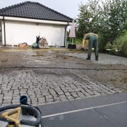 Mikołaj Nowak - Czyszczenie kostki brukowej na posesji. Mężczyzna zamiata mokrą nawierzchnię. W tle budynek z ciemnym dachem i zieleń. Widoczny wąż ogrodowy i butla gazowa.