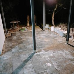 Mikołaj Nowak - Ułożona szara kostka brukowa na zewnątrz pod zadaszeniem, widoczne białe wiadra i narzędzia. Praca w toku, oświetlenie nocne.