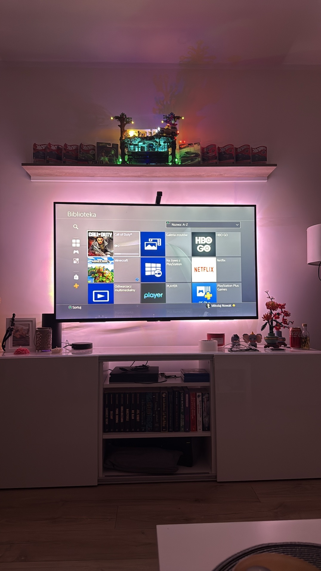 Telewizor z podświetleniem LED na szafce RTV, nad nim półka z dekoracjami i zabawkami. Całość w stonowanej kolorystyce, oświetlona ciepłym światłem.