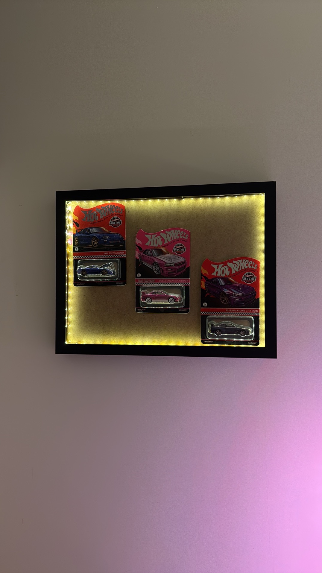 Podświetlana rama z kolekcją autek Hot Wheels Red Line: Toyota Supra i Nissan Skyline GT-R (R34) w oryginalnych opakowaniach, zawieszona na ścianie.
