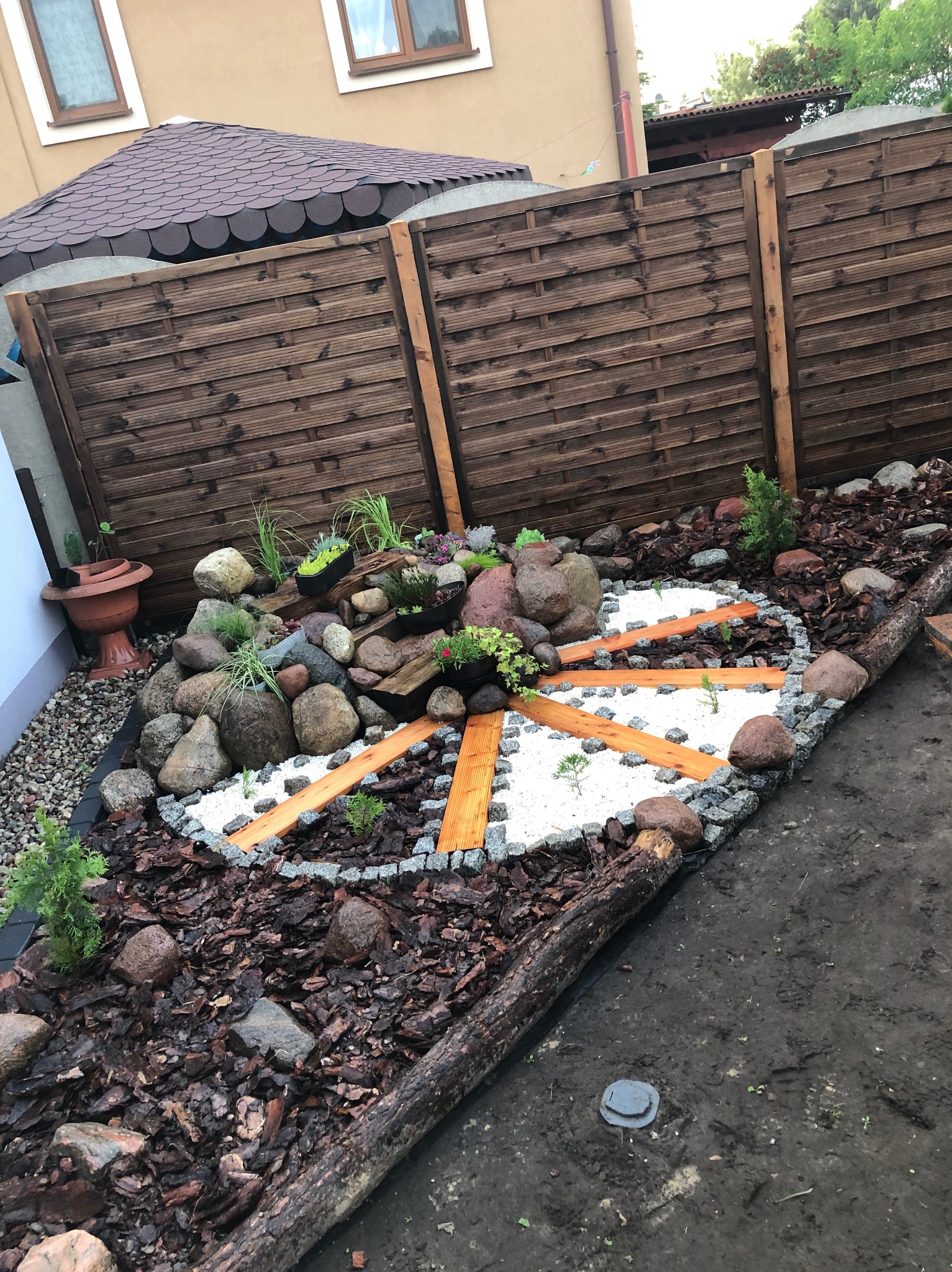 Ogród z dekoracyjnym układem kamieni, drewna i białego żwiru, tworzącym wzór koła z promienistymi ścieżkami. Tło stanowi drewniany płot i fragment budynku.