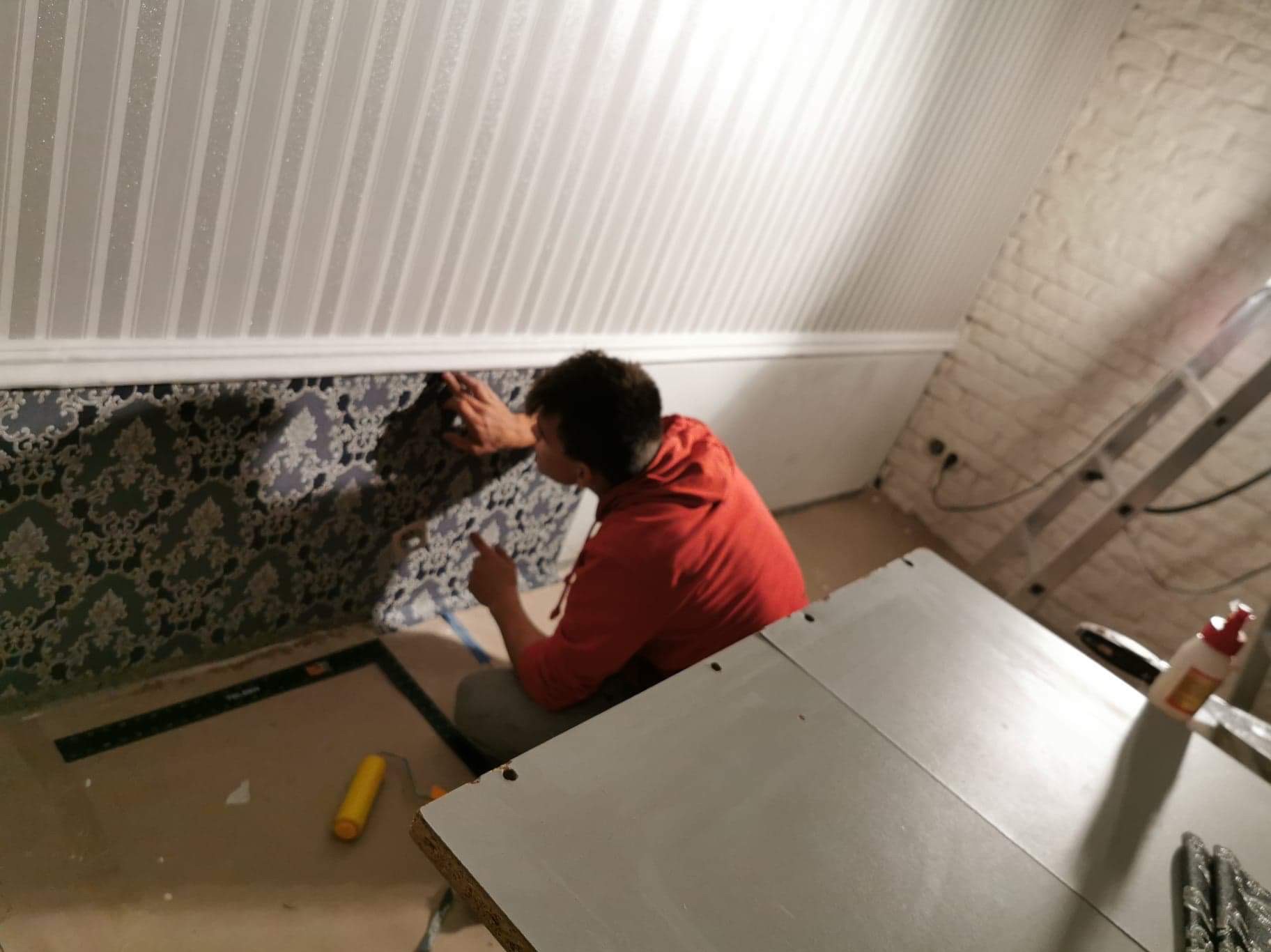 Pracownik w czerwonej bluzie przykleja wzorzystą tapetę na ścianie pod skośnym sufitem. Widoczny wałek malarski i stół roboczy. Część ściany wykończona w białe pasy.