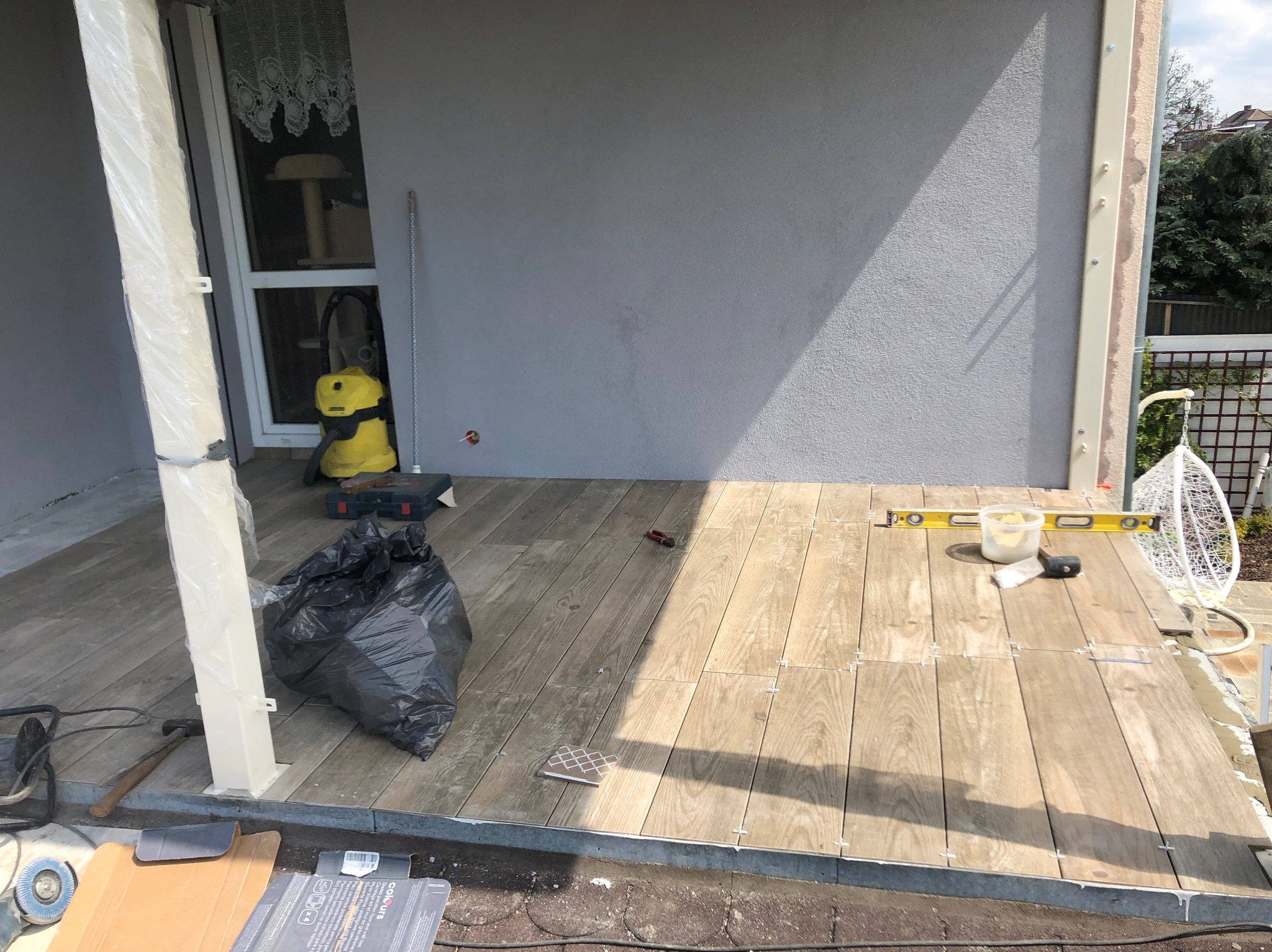 Układanie drewnopodobnych płytek na tarasie. Widoczne poziomica, wiadro z zaprawą i narzędzia. Częściowo ułożona podłoga z płytek. Na zewnątrz, w słoneczny dzień.