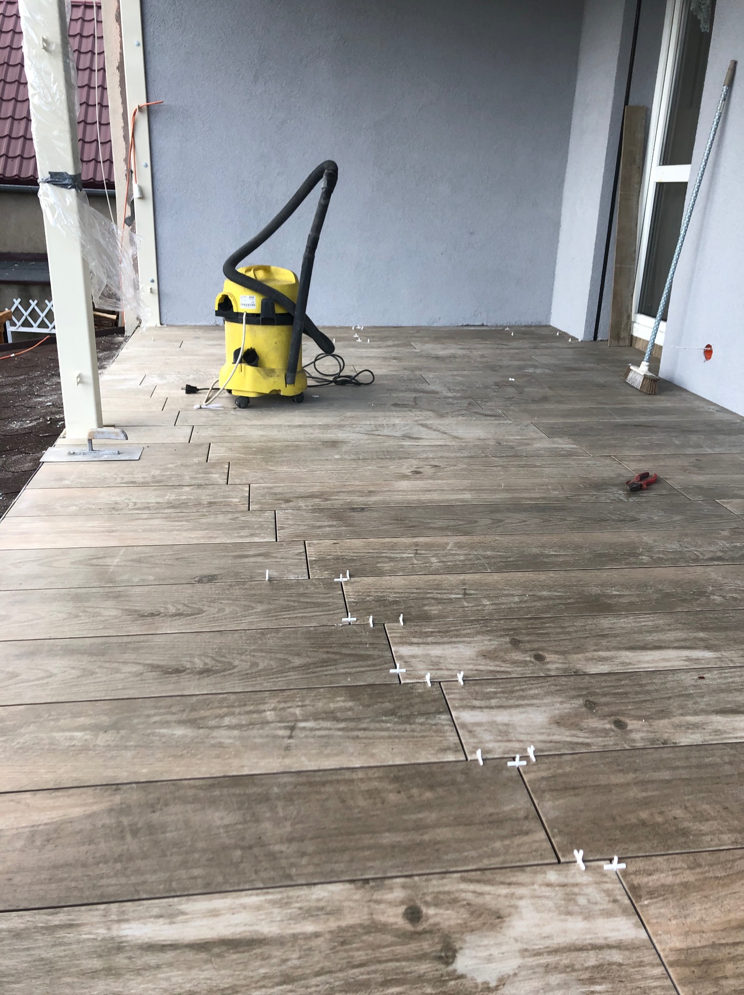 Układanie drewnopodobnych płytek na tarasie. Widoczny odkurzacz przemysłowy, krzyżyki dystansowe i szczotka. Częściowo ułożona podłoga na zewnątrz budynku.
