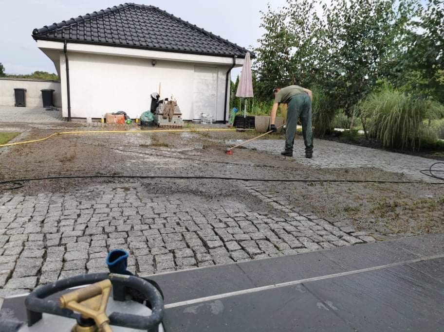 Czyszczenie kostki brukowej na posesji. Mężczyzna zamiata mokrą nawierzchnię. W tle budynek z ciemnym dachem i zieleń. Widoczny wąż ogrodowy i butla gazowa.