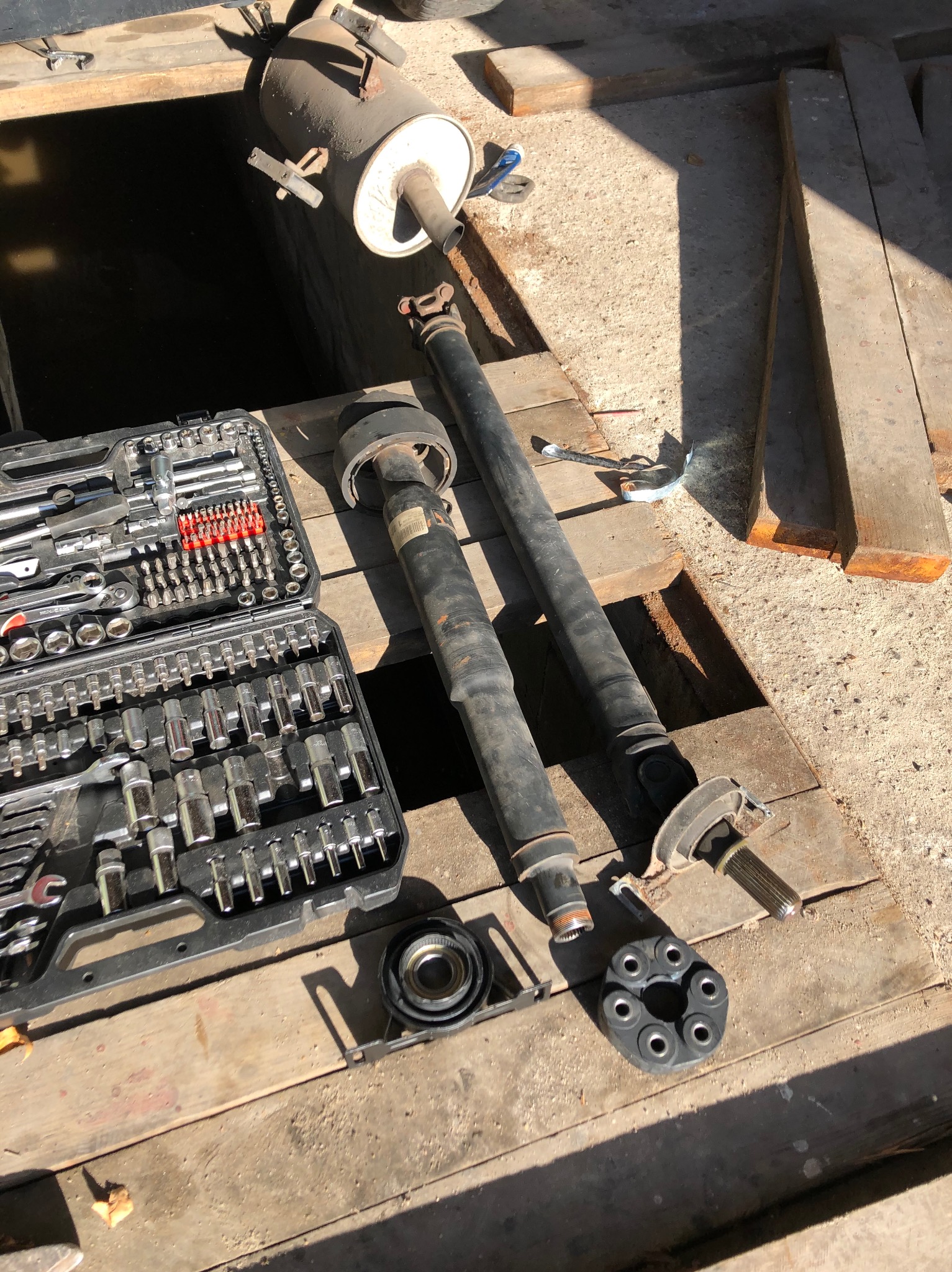 Warsztatowy nieład: skrzynka narzędziowa, wały napędowe i tłumik na drewnianej podłodze garażu z betonową posadzką. Widoczny otwór w podłodze.