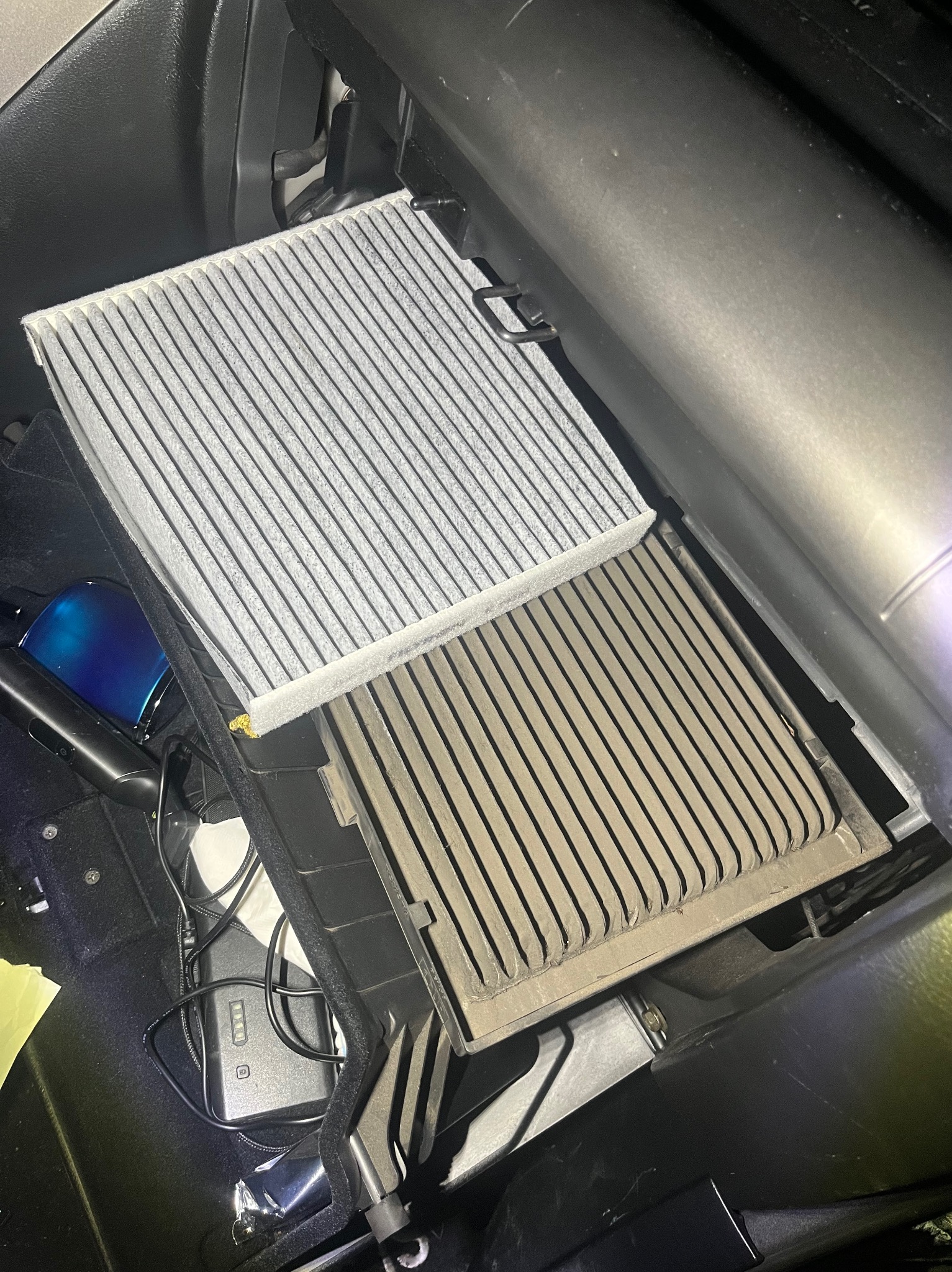 Wymiana filtra kabinowego w samochodzie: nowy, biały filtr obok starego, szarego. Widoczne wnętrze schowka i akcesoria.