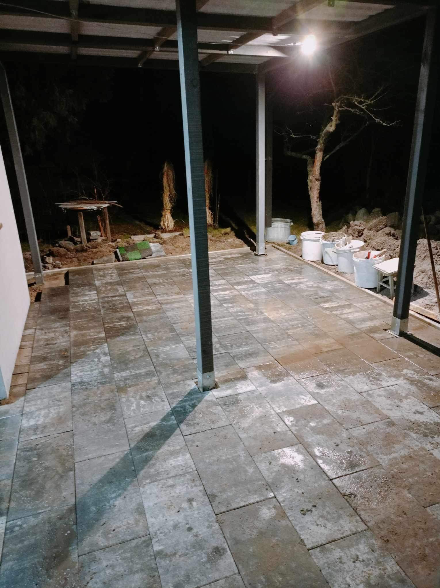 Ułożona szara kostka brukowa na zewnątrz pod zadaszeniem, widoczne białe wiadra i narzędzia. Praca w toku, oświetlenie nocne.
