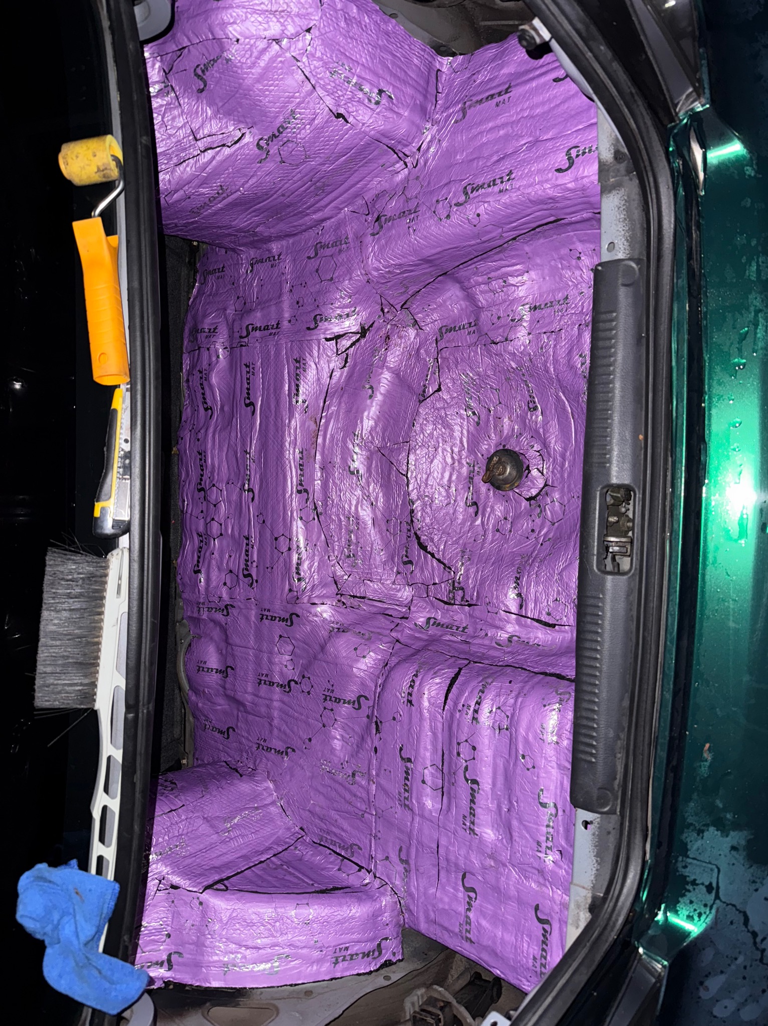 Fioletowa mata wygłuszająca Smart Mat w bagażniku samochodu. Widoczny wałek malarski, nóż i szczotka. Wygłuszenie bagażnika w trakcie prac.