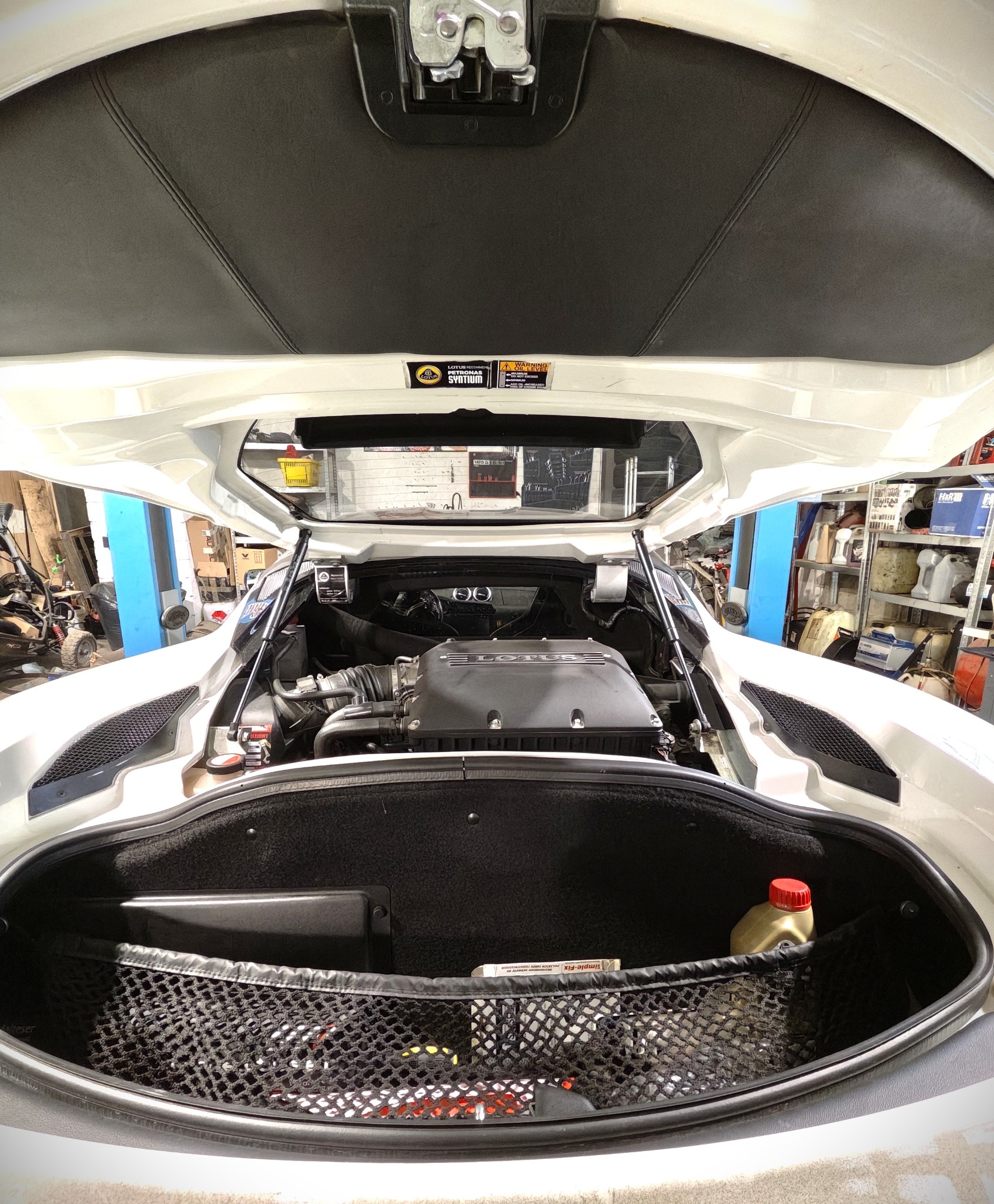 Otwarta klapa bagażnika białego Lotus'a z widocznym silnikiem i elementami wyposażenia warsztatu w tle. Widok z góry, wnętrze bagażnika z siatką i butelką oleju.