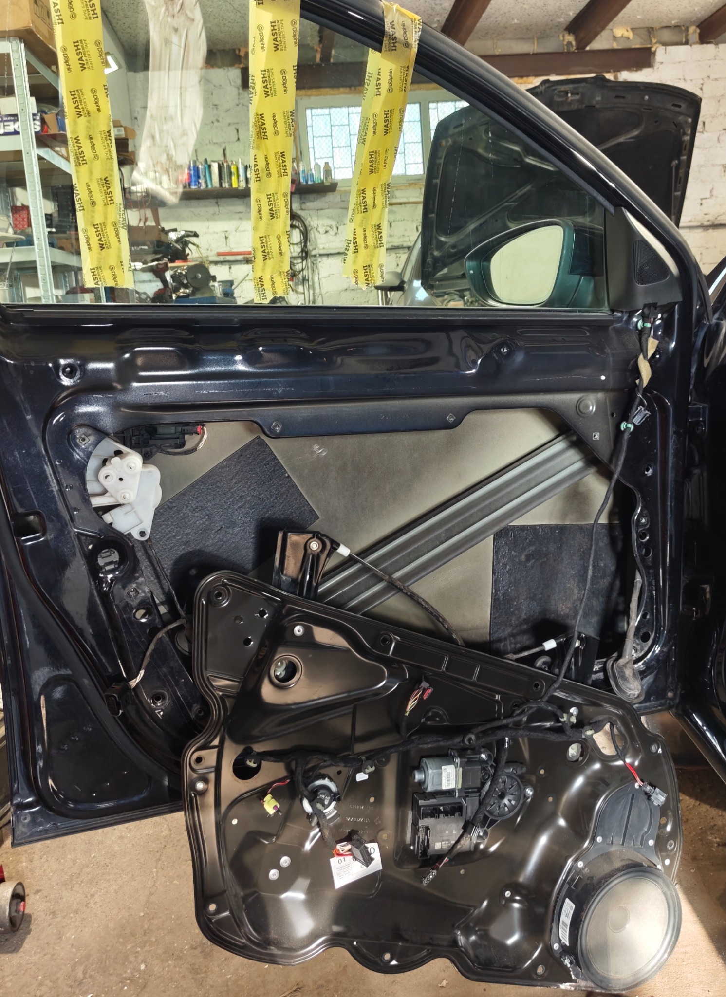 Demontaż tapicerki drzwi auta w warsztacie. Widoczne elementy mechanizmu okna i głośnik. Żółta taśma malarska zabezpiecza szybę. Otwarta maska w tle.