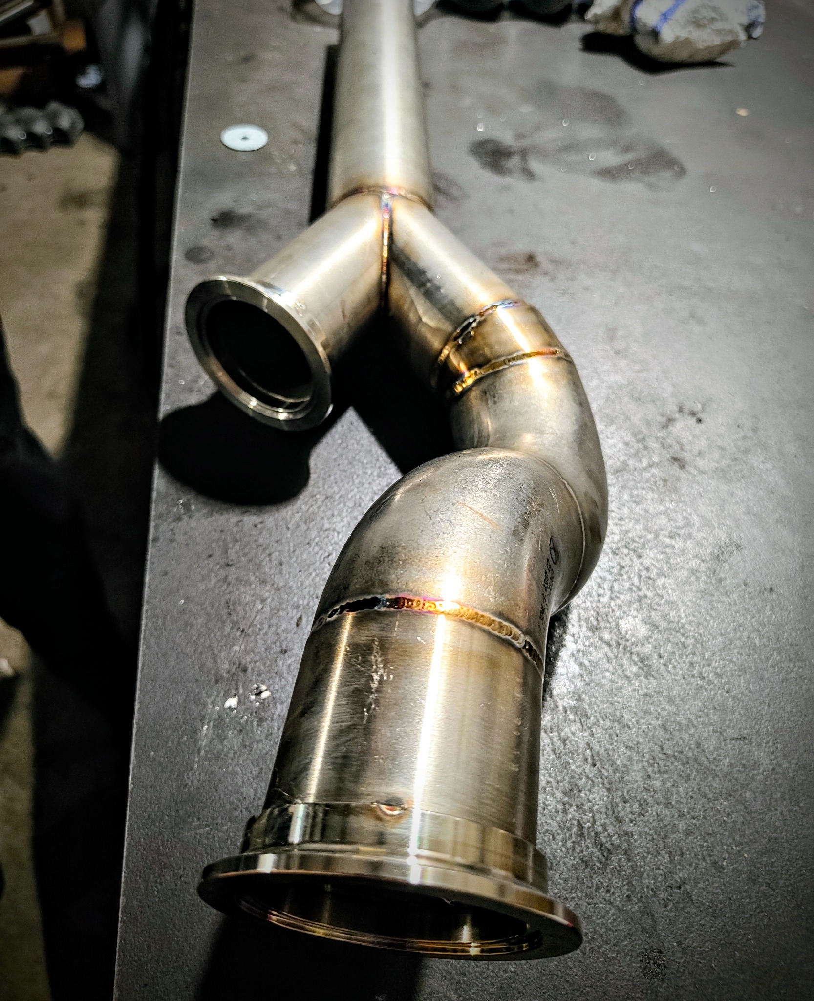 Spawany, nierdzewny układ wydechowy w kształcie litery Y, leżący na szarym stole warsztatowym. Widoczne spawy i metaliczne wykończenie.