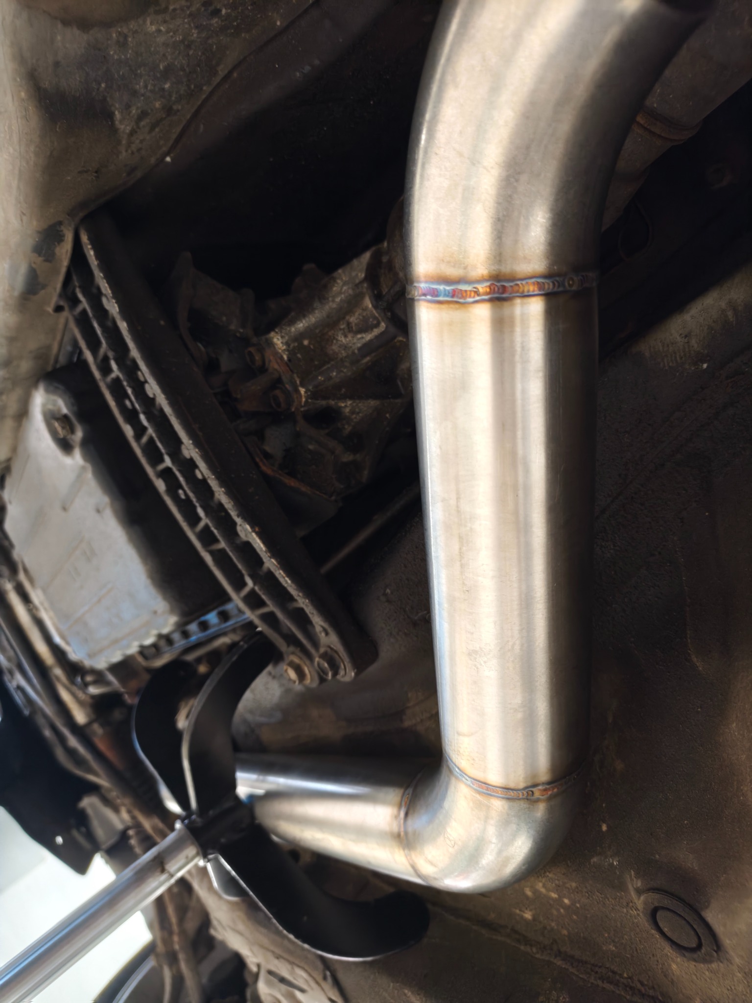 Spawany, nierdzewny układ wydechowy pod podwoziem auta, widoczne spawy z charakterystycznymi przebarwieniami termicznymi. Elementy zawieszenia i skrzyni biegów w tle.