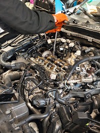 Silnik samochodowy w trakcie naprawy: widoczne elementy wewnętrzne, ręka w pomarańczowej rękawicy używa narzędzia. Telefon leży obok, oświetlenie warsztatowe.