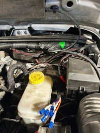 Otwarta maska samochodu z widocznymi przewodami, zbiornikiem płynu hamulcowego z żółtą nakrętką i złączkami elektrycznymi. Detal podzespołów w komorze silnika.