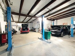 Wnętrze warsztatu samochodowego z dwoma podnośnikami, czerwonym autem z otwartym bagażnikiem i czarnym autem z podniesioną maską. Widoczne narzędzia i wyposażenie warsztatowe.