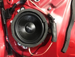 Głośnik STEG zamontowany w czerwonych drzwiach samochodu, widoczne przewody i elementy montażowe. Detal instalacji audio w aucie, z bliska.